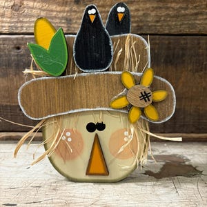 May include: Wooden scarecrow decoration with a straw hat, featuring two black bird figures, a yellow flower, and corn stalks. The scarecrow has a painted face with rosy cheeks and an orange triangle nose. The hat is trimmed with white paint.