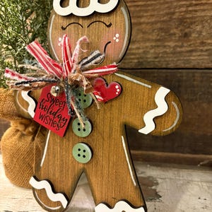 May include: Wooden gingerbread man decoration with white accents and a red tag that says "Sweet holiday wishes." The gingerbread man has a white hat, green buttons, and a red heart. A small burlap sack is visible in the background.