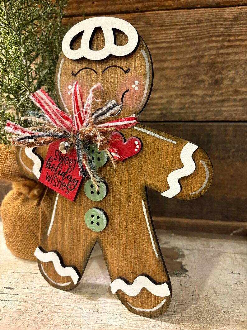 May include: Wooden gingerbread man decoration with white accents and a red tag that says "Sweet holiday wishes." The gingerbread man has a white hat, green buttons, and a red heart. A small burlap sack is visible in the background.