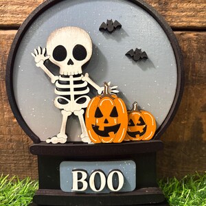 May include: A Halloween-themed wooden decoration featuring a skeleton waving, two orange jack-o'-lanterns, and two black bats against a light blue background. The word "BOO" is displayed on a blue plaque.