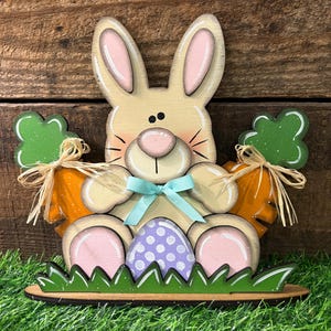 May include: Wooden Easter bunny decoration with a light tan body, pink accents, and a blue bow. It holds two orange carrots with green tops and a purple polka-dotted egg. The bunny sits on a green grass base.