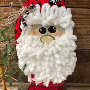 May include: A whimsical Santa Claus decoration with a red hat adorned with black polka dots and a white snowflake. The face features large, expressive eyes and a fluffy white beard. The base is red.