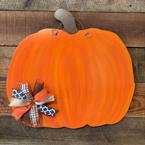 May include: A large, orange, wooden pumpkin decoration with a metal stem and a decorative bow. The bow features ribbons in shades of orange, brown, white, and navy blue. The pumpkin is painted with a gradient effect, and has two small holes for hanging.