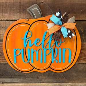 May include: Orange wooden pumpkin-shaped sign with the words "hello PUMPKIN" in turquoise. The sign has a black outline and a decorative bow with turquoise, orange, and brown ribbons. A brown stem and a wire hanger complete the design.
