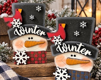 May include: Two wooden snowman decorations featuring black top hats with the word Winter written in white script. Each snowman has a carrot nose, a red cardinal, white snowflake cutouts, and a plaid scarf in either red or blue patterns.