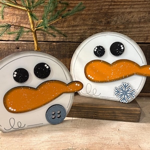 Op de afbeelding: Twee houten sneeuwmanversieringen. Elke sneeuwman heeft een wit gezicht, zwarte ogen en een oranje wortelneus. Eén heeft een blauwe knoop en de andere een sneeuwvlok. De sneeuwmannen staan op een houten oppervlak.