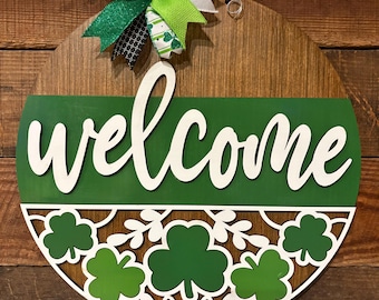 May include: A circular wooden welcome sign featuring a green and white shamrock design. The word "welcome" is in white script across a green section. A decorative bow with green and white ribbons adorns the top.