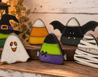 Puede incluir: Un conjunto de seis decoraciones de madera para Halloween con forma de caramelos de maíz, que incluye una bruja, un fantasma, un caramelo clásico, una figura inspirada en Frankenstein, un murciélago y una momia en colores vibrantes.