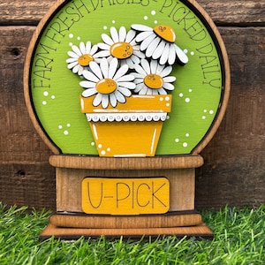 May include: A wooden sign with a green background and a daisy flower arrangement in a yellow pot. The sign reads "Farm Fresh Daisies - Picked Daily" and "U-Pick". The sign is on a wooden base.