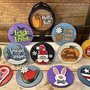 May include: A collection of 12 round wooden signs with various seasonal and holiday themes. The signs feature painted designs and text, including "Trick or Treat", "Hello Fall", "Fresh Picked Daisies", "Freezin Season", "Give Thanks", "Back to School", "Top 14", and more.