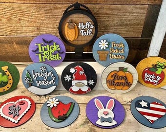 Può includere: Una collezione di 12 cartelli rotondi in legno con vari temi stagionali e festivi. I cartelli presentano disegni dipinti e testo, tra cui "Trick or Treat", "Hello Fall", "Fresh Picked Daisies", "Freezin Season", "Give Thanks", "Back to School", "Top 14", e altro ancora.