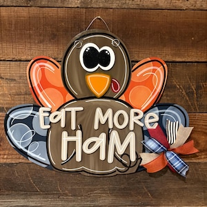 May include: A wooden turkey-shaped sign with the text "EAT MORE HAM". The turkey has brown, orange, and blue accents, with a decorative bow. The sign is designed for Thanksgiving or fall decor.