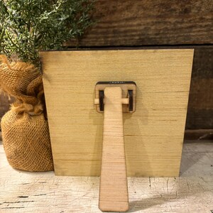 May include: A square light-colored wooden plaque featuring a hinged easel stand attached to the back center. The plaque is constructed from smooth, flat wood with a visible grain pattern and a dark brown edge, designed for tabletop display.