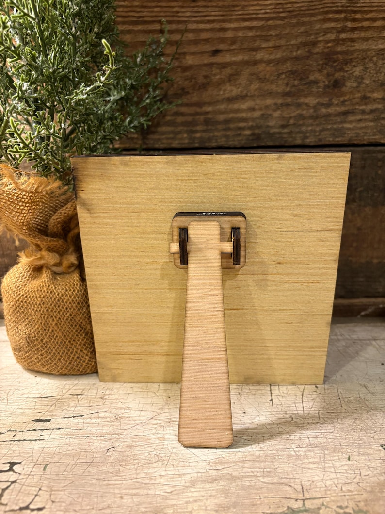 May include: A square light-colored wooden plaque featuring a hinged easel stand attached to the back center. The plaque is constructed from smooth, flat wood with a visible grain pattern and a dark brown edge, designed for tabletop display.