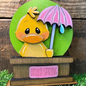 May include: A wooden spring-themed decoration featuring a yellow duck with an orange beak and a pink umbrella. The duck is set against a green backdrop, and the base has a pink sign that reads "HELLO SPRING".