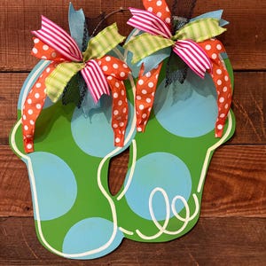 May include: A pair of wooden flip-flops painted green with blue polka dots and a blue background. The flip-flops are adorned with orange and white polka dot ribbons, pink and white striped ribbons, and green striped ribbons. Black mesh and blue ribbon complete the design.