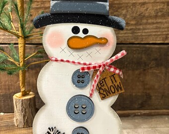 May include: Wooden snowman decoration with a black top hat, orange carrot nose, and blue button details. The snowman has a red and white checkered scarf and a wooden tag that says "Let It Snow".