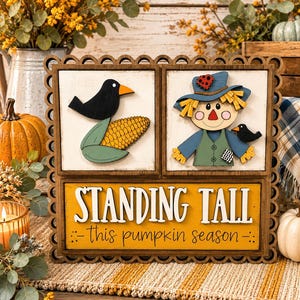 May include: A wooden decorative sign with a scalloped border featuring three sections. The top left shows a black crow and corn, the top right a scarecrow with a crow, and the bottom yellow panel reads STANDING TALL this pumpkin season.