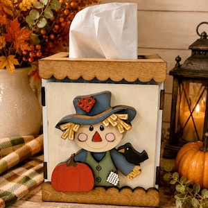 May include: A square wooden tissue box cover featuring a layered cutout of a scarecrow wearing a blue hat with a red patch, a green vest, and a black crow. The scarecrow holds a large orange pumpkin. The box has scalloped top and bottom edges.