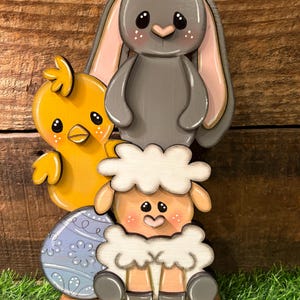 May include: A wooden Easter decoration featuring a gray bunny with pink inner ears, a yellow chick, a sheep, and a blue egg with white floral designs. The figures are painted with soft colors and sit on a wooden base, perfect for spring decor.