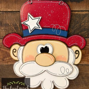 May include: A wooden patriotic door hanger shaped like a face with a red top hat, blue band, and a white star. The face has a tan complexion, a white beard, and a tan nose. The text "the front porch southern decor" is in the bottom left corner.