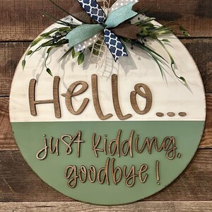 Puede incluir: Un letrero redondo de madera con el texto "Hello... just kidding, goodbye!" en una fuente juguetona. La mitad superior es blanca, la inferior es verde, decorado con un lazo y vegetación.