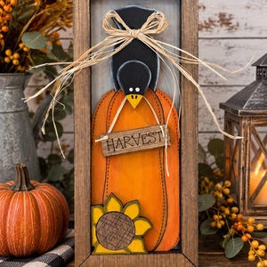 May include: A rectangular wooden framed sign featuring a layered cutout of an orange pumpkin, a black crow, and a yellow sunflower. A small wooden plaque hanging from the crow's beak displays the word HARVEST in black text, accented by a raffia bow.