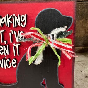 May include: A rectangular red wooden sign featuring a black silhouette of a dog wearing a frayed fabric bow in red, white, and green. The sign includes raised white lettering with black outlines that partially reads, "making it, I've seen it twice."