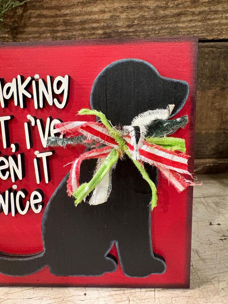 May include: A rectangular red wooden sign featuring a black silhouette of a dog wearing a frayed fabric bow in red, white, and green. The sign includes raised white lettering with black outlines that partially reads, "making it, I've seen it twice."