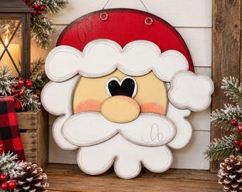 May include: A round wooden Santa Claus wall hanging featuring a red hat with white trim, a pale yellow face with rosy cheeks, and a large white mustache. The piece includes a metal wire hanger at the top and has a smooth, painted finish.