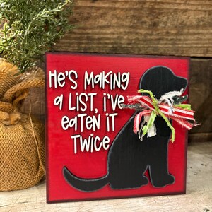 May include: A square red wooden sign featuring a black silhouette of a dog wearing a frayed red, white, and green ribbon around its neck. The sign displays the white text: He's Making a LiST, i've eaTeN iT Twice. The edges of the sign are dark.