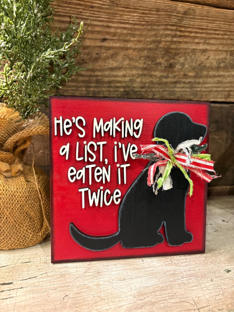 May include: A square red wooden sign featuring a black silhouette of a dog wearing a frayed red, white, and green ribbon around its neck. The sign displays the white text: He's Making a LiST, i've eaTeN iT Twice. The edges of the sign are dark.