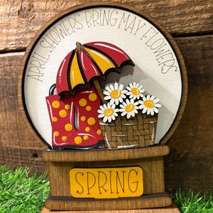 May include: Wooden spring-themed decoration featuring a red and yellow umbrella, red rain boots with yellow polka dots, and a basket of daisies. The text "APRIL SHOWERS BRING MAY FLOWERS" is written around the top, and "SPRING" is written on the base.