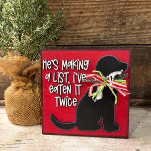 May include: A square red wooden sign featuring a black silhouette of a dog wearing a frayed red, white, and green ribbon around its neck. The text reads, "He's Making a LIST, i've eaTen iT Twice" in white lettering with a black outline.