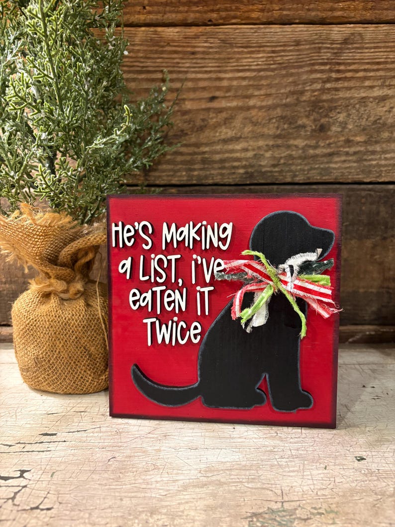 May include: A square red wooden sign featuring a black silhouette of a dog wearing a frayed red, white, and green ribbon around its neck. The text reads, "He's Making a LIST, i've eaTen iT Twice" in white lettering with a black outline.