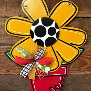 May include: A yellow flower with a black and white polka dot center, in a red pot with a green stem. The flower has a yellow and red bow with a white polka dot ribbon.