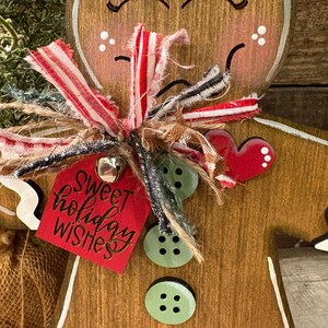May include: Wooden gingerbread man Christmas ornament with a red tag that says "Sweet holiday wishes". The ornament has a white painted outline, a red heart, and green button accents. It is decorated with a red and white striped ribbon.
