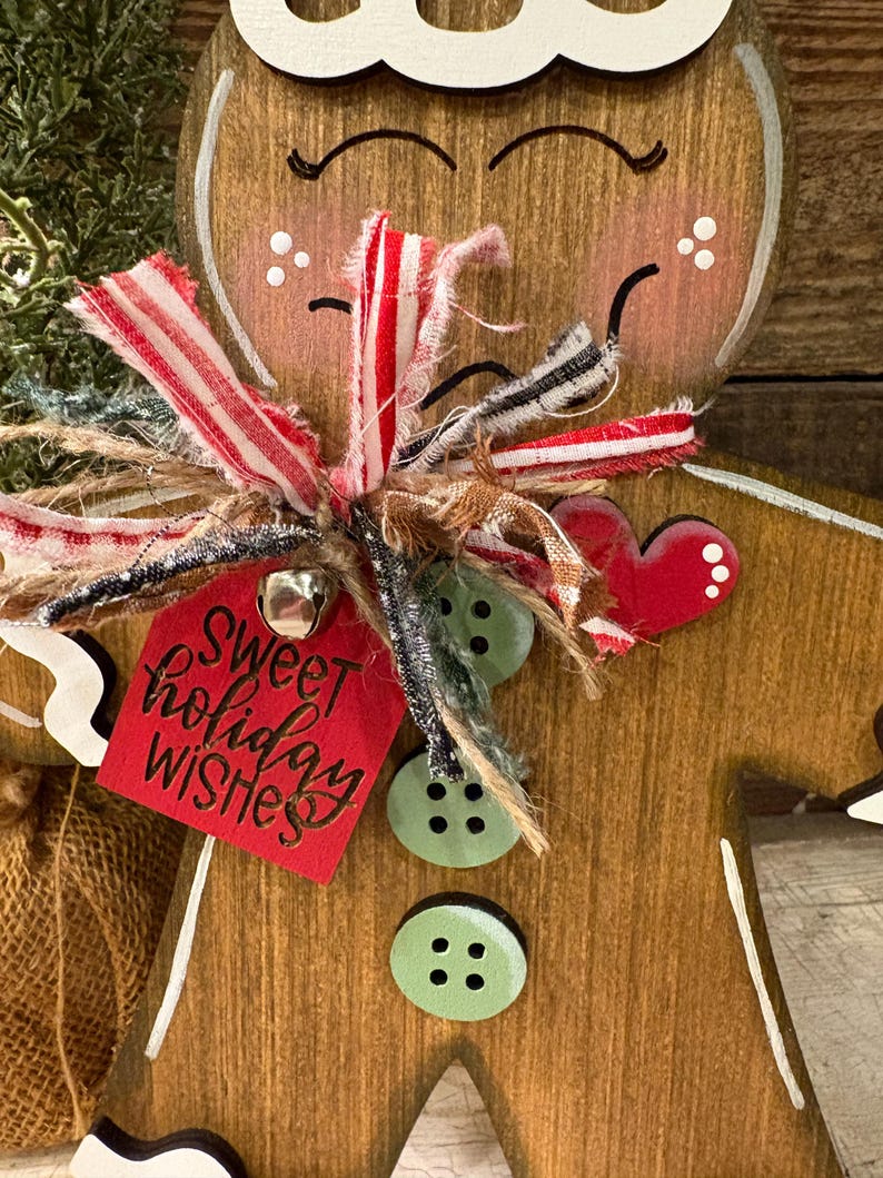 May include: Wooden gingerbread man Christmas ornament with a red tag that says "Sweet holiday wishes". The ornament has a white painted outline, a red heart, and green button accents. It is decorated with a red and white striped ribbon.