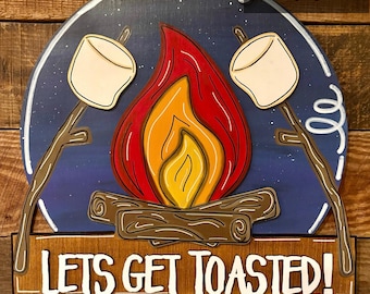 Könnte beinhalten: Ein rundes Holzschild mit blauem Hintergrund und einer Feuerstellenszene. Das Lagerfeuer hat orange und gelbe Flammen und zwei weiße Marshmallows auf Stöcken. Das Schild trägt den Text "LETS GET TOASTED!" in weißen Buchstaben.