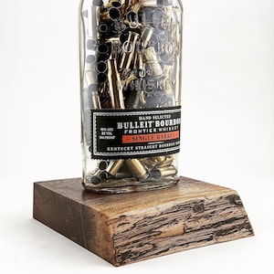 May include: A unique lamp made from a Bulleit Bourbon bottle filled with brass bullet casings. The bottle sits on a wooden base. The lamp has a white lampshade and a cork stopper.