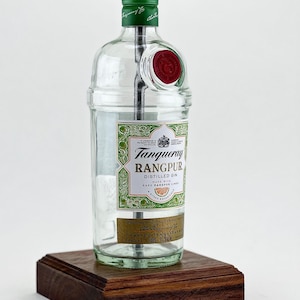 May include: A clear glass bottle lamp with a white lampshade. The bottle is labeled "Tanqueray Rangpur Distilled Gin" with a green and gold design. The bottle is sitting on a dark wood base.