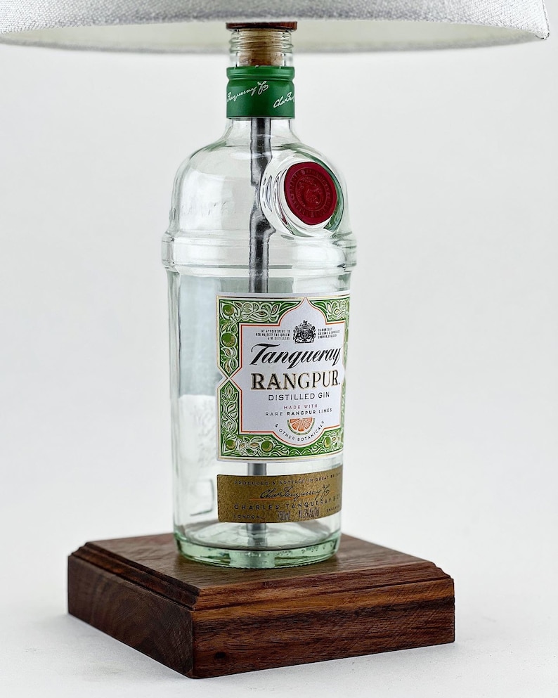 May include: A clear glass bottle lamp with a white lampshade. The bottle is labeled "Tanqueray Rangpur Distilled Gin" with a green and gold design. The bottle is sitting on a dark wood base.