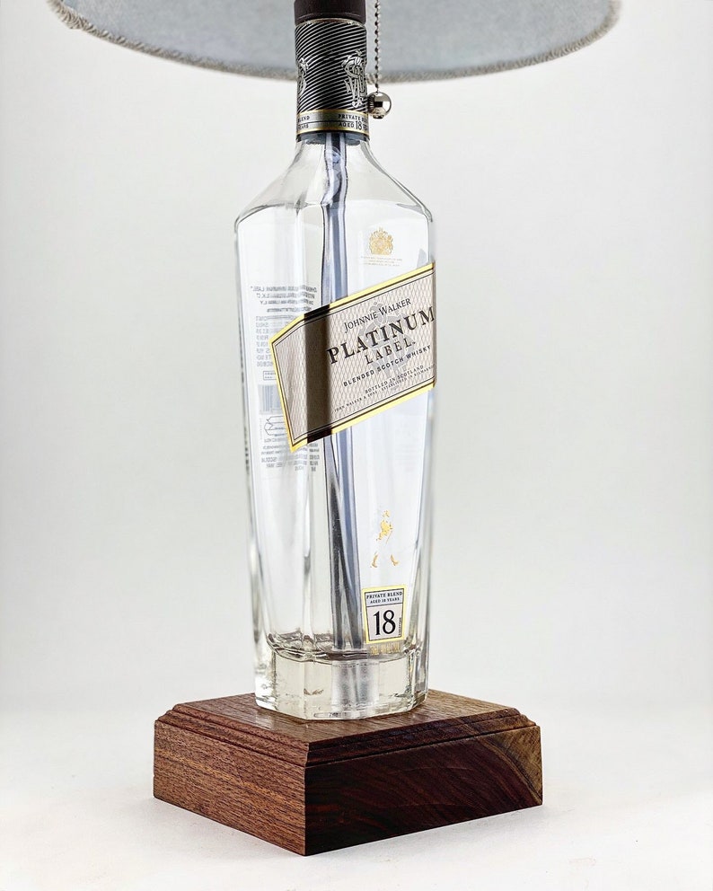 Może przedstawiać: Lampka z przezroczystej szklanej butelki z drewnianą podstawą. Butelka jest oznaczona etykietą "Johnnie Walker Platinum Label Blended Scotch Whisky". Butelka ma srebrną metalową zakrętkę i srebrny łańcuszek z wiszącą srebrną kulką. Klosz lampy jest jasnoszary.