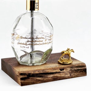 May include: A clear glass bottle lamp with a gold top and a white lampshade. The bottle has gold lettering that reads "Bourbon whiskey dumped on Warehouse Stored in Individually selected filtered and bottled by hand 51.5% vol". The lamp sits on a wooden base with a gold horse figurine.