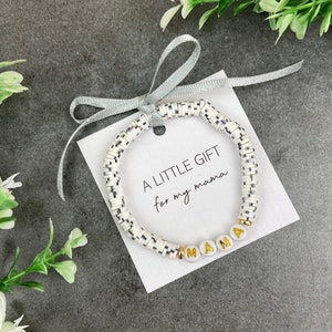 Op de afbeelding: Een wit en zwart kralenarmband met gouden letterkralen die "MAMA" spellen. De armband is vastgebonden met een zilveren lint en een witte kaart waarop staat "A LITTLE GIFT for my mama".