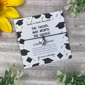 Op de afbeelding: Witte kaart met zwarte afstudeerhoeden erop gedrukt. De kaart heeft de tekst "Make a Wish Bracelet" en "The tassel was worth the hassle". De kaart heeft ook de tekst "Congratulations! Close your eyes and make a wish, then tie this bracelet on your wrist. When it falls off or wears through, that is when your wish comes true." en "The Persnickety Co."