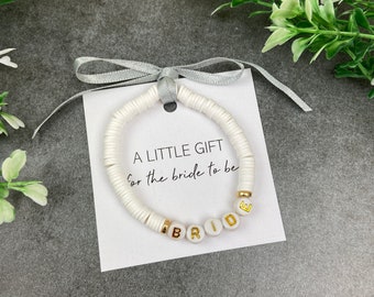 May include: White beaded bracelet with the word "BRIDE" spelled out in gold letters. The bracelet is on a white card with the text "A LITTLE GIFT for the bride to be".