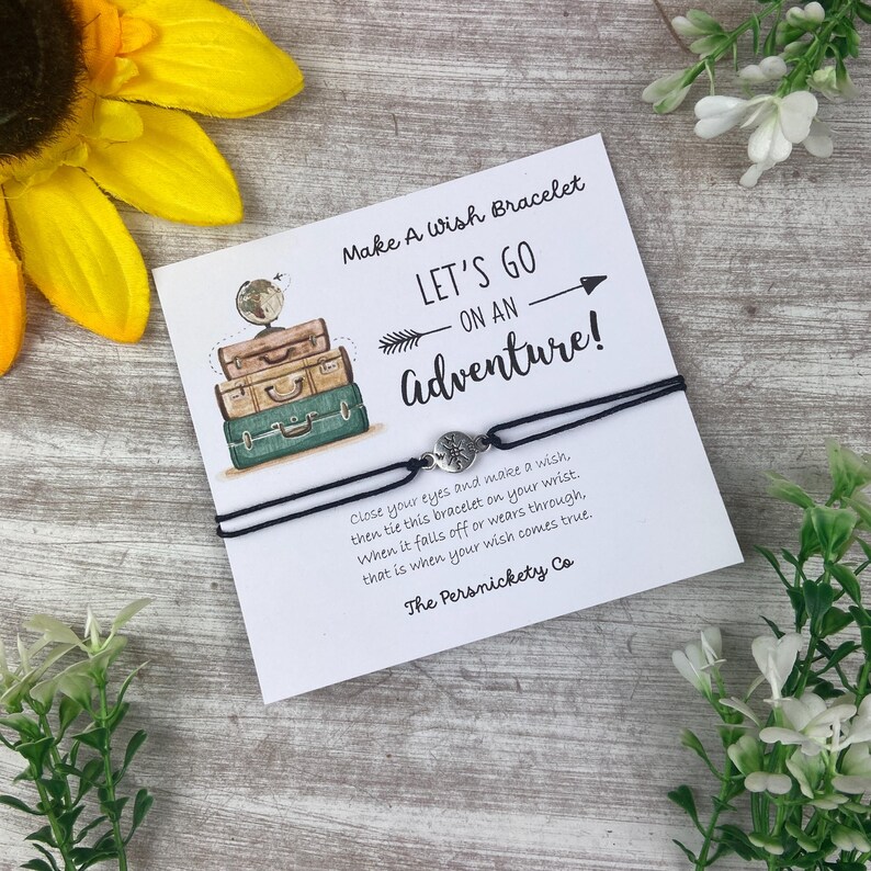 May include: A black string bracelet with a compass charm is displayed on a card that reads "Make A Wish Bracelet" and "Let's Go on an Adventure!". The card also features a stack of vintage suitcases.