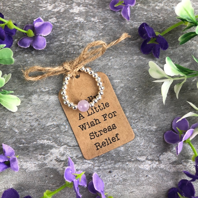May include: A silver beaded ring with a light purple stone. The ring is on a brown tag with the text "A Little Wish For Stress Relief".