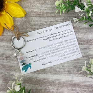 Puede incluir: Una tarjeta blanca con el texto "Safe Travels - Travel Turtle" y una descripción del significado de diferentes piedras preciosas. La tarjeta está junto a un llavero plateado con un pequeño amuleto de tortuga y una pila de piedras de colores.